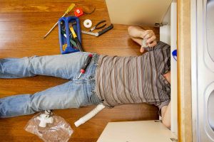 A man on the kitchen floor is fixing the sink.
